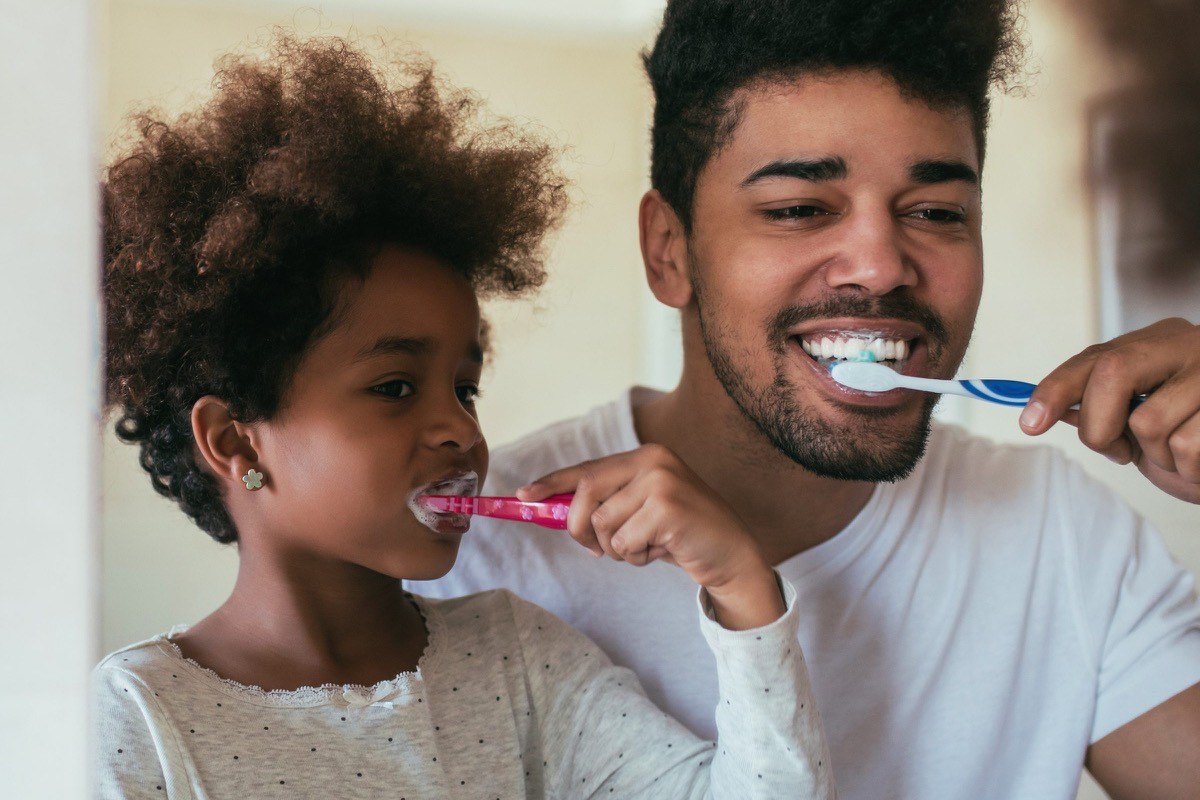 A father and his young daughter brushing their teeth together at the bathroom mirror