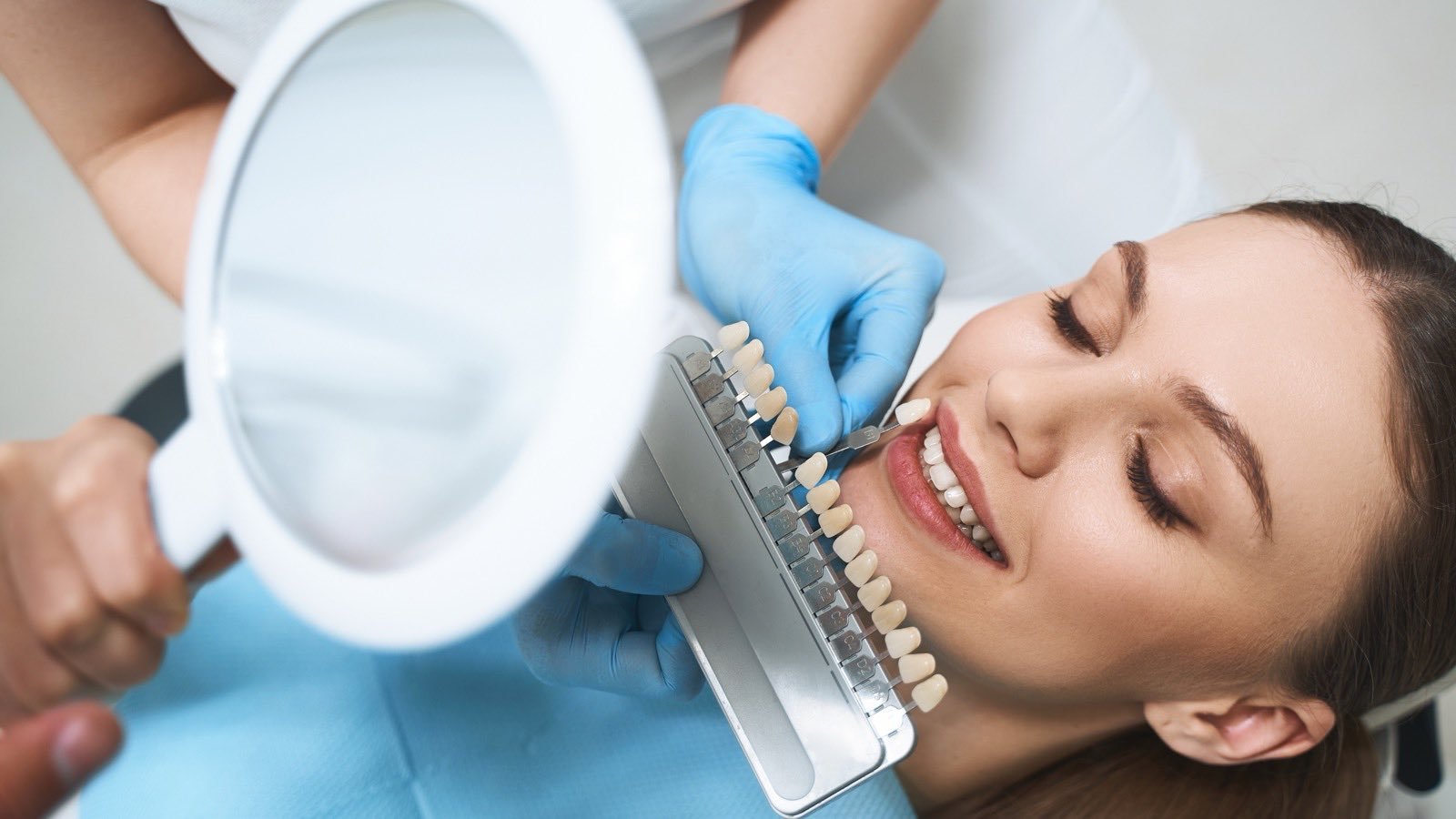 A dentist matching a restoration shade against a patient’s teeth while she checks the result in a hand mirror